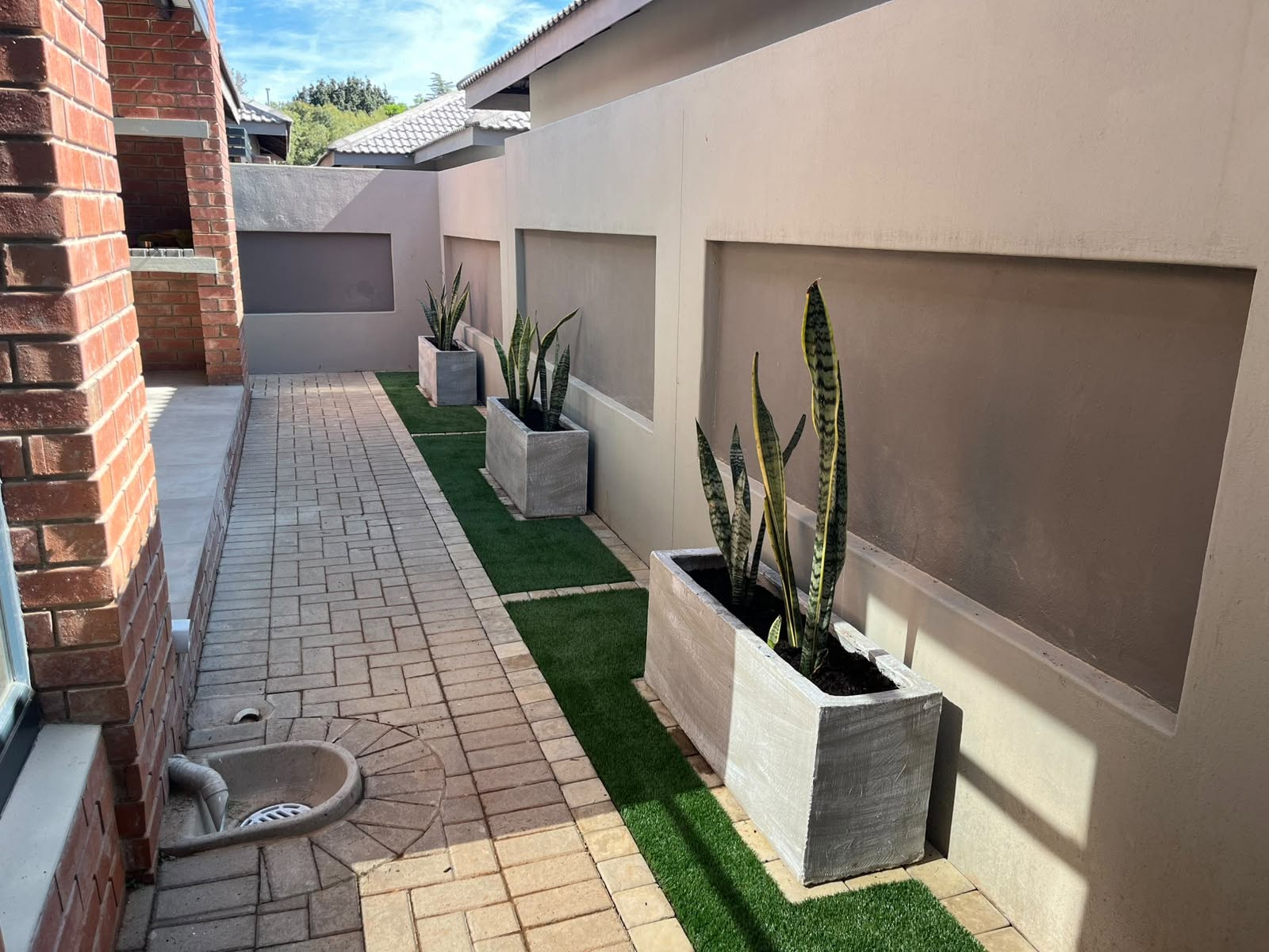 Narrow side walkway with artificial grass strips and raised planters against a brick wall.