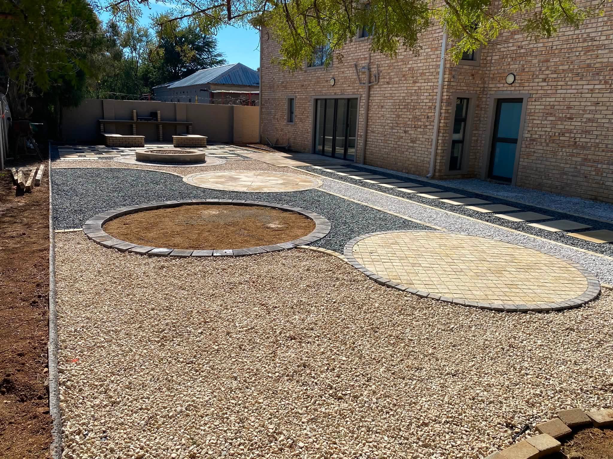 Circular and oval paving set into a gravel courtyard with a central fire-pit ring, next to a face-brick building.