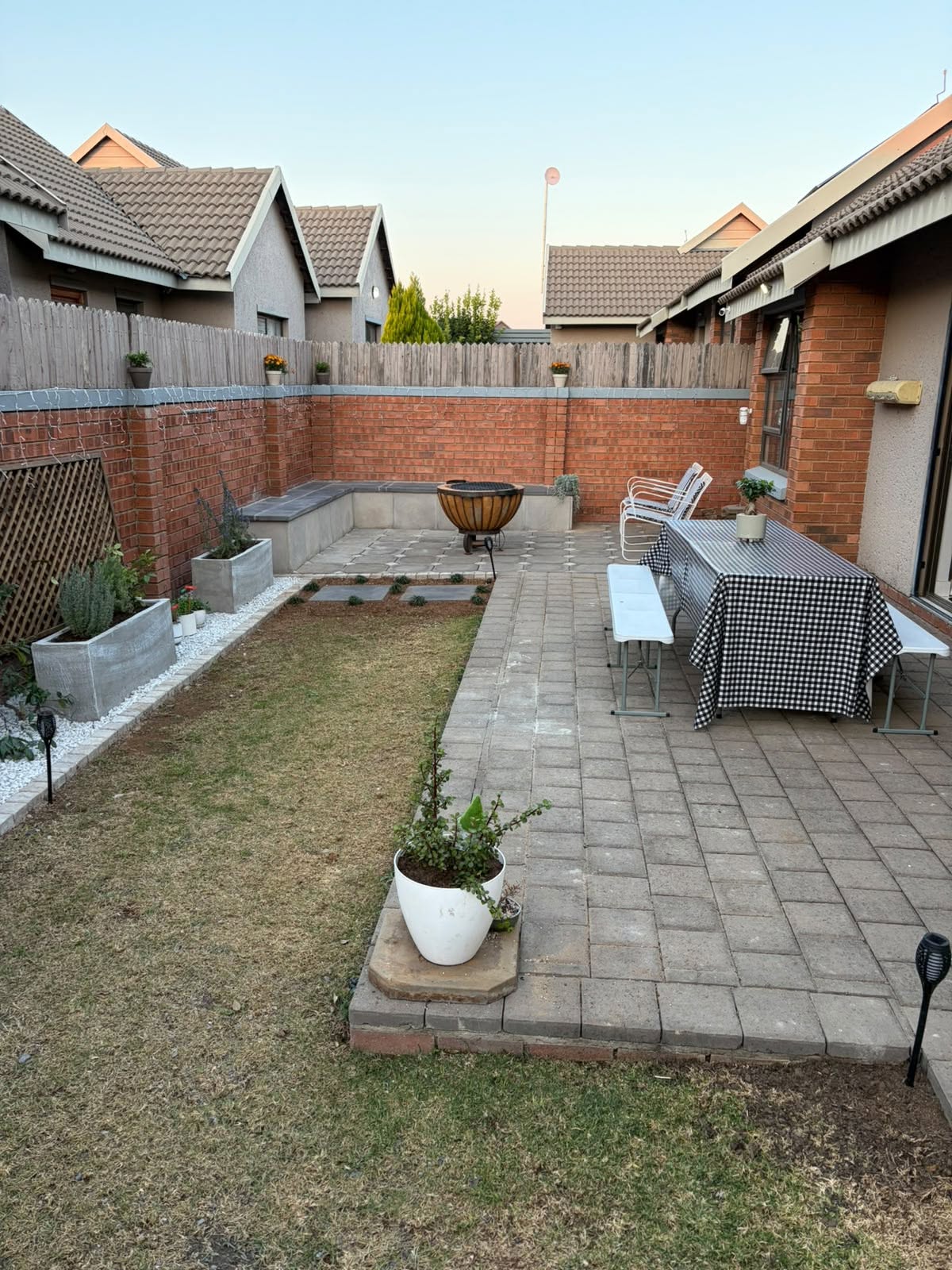Paved patio with built-in bench seating, a dining table set for a gathering and lattice screening, backed by a red-brick boundary wall.