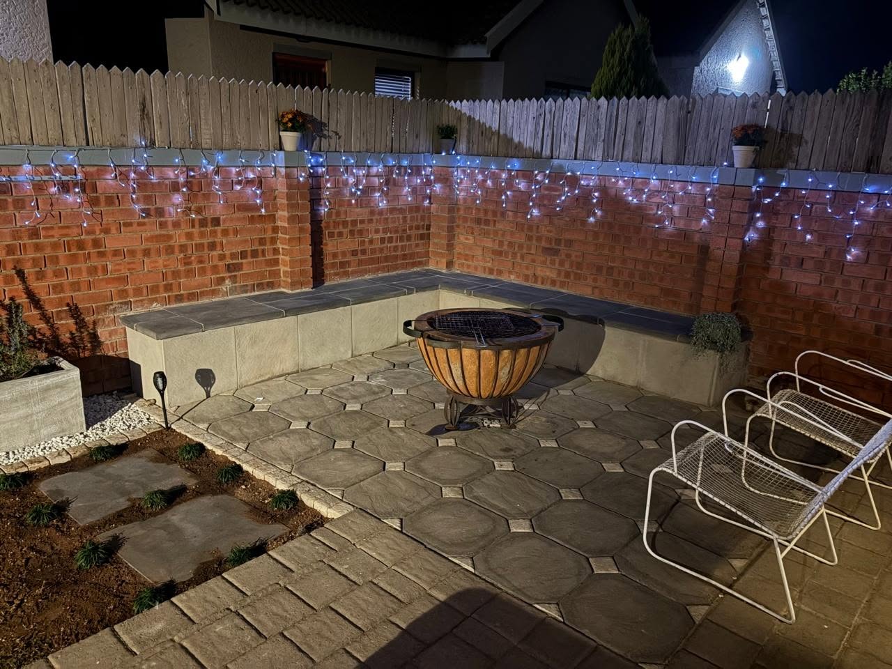 Patio and built-in seating area lit at night around a fire pit, with festoon lighting along the brick boundary wall.